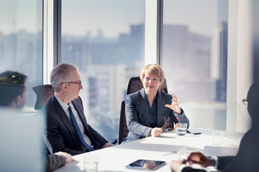 Female in a business suit talking to peers while holding reading glasses in her one hand.